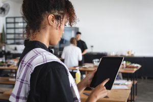 A Black woman taps on a tablet while inside a restaurant kitchen.