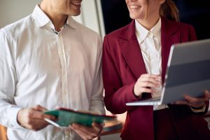 A man and women in business professional clothes smile at each other. The woman has an open laptop in her hands.