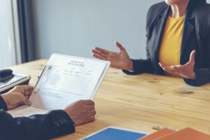 Picture of two people sitting at a table and talking while one holds a clipboard.